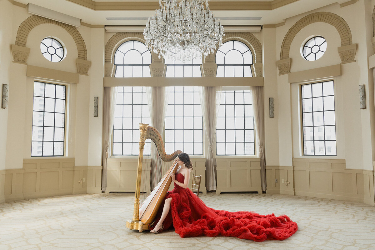 woman in long red gown is sitting at her harp in a formal room with very tall windows and chandeleir.
