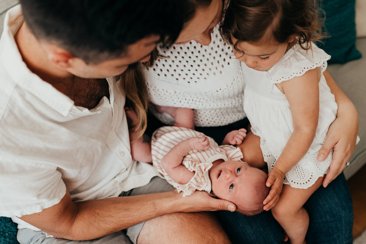 A mother and father hold their older daughter and their newborn daughter on their couch, the big sister gently touches her sister's head during her photoshoot with Allison Wolf, Boston Newborn Photographer