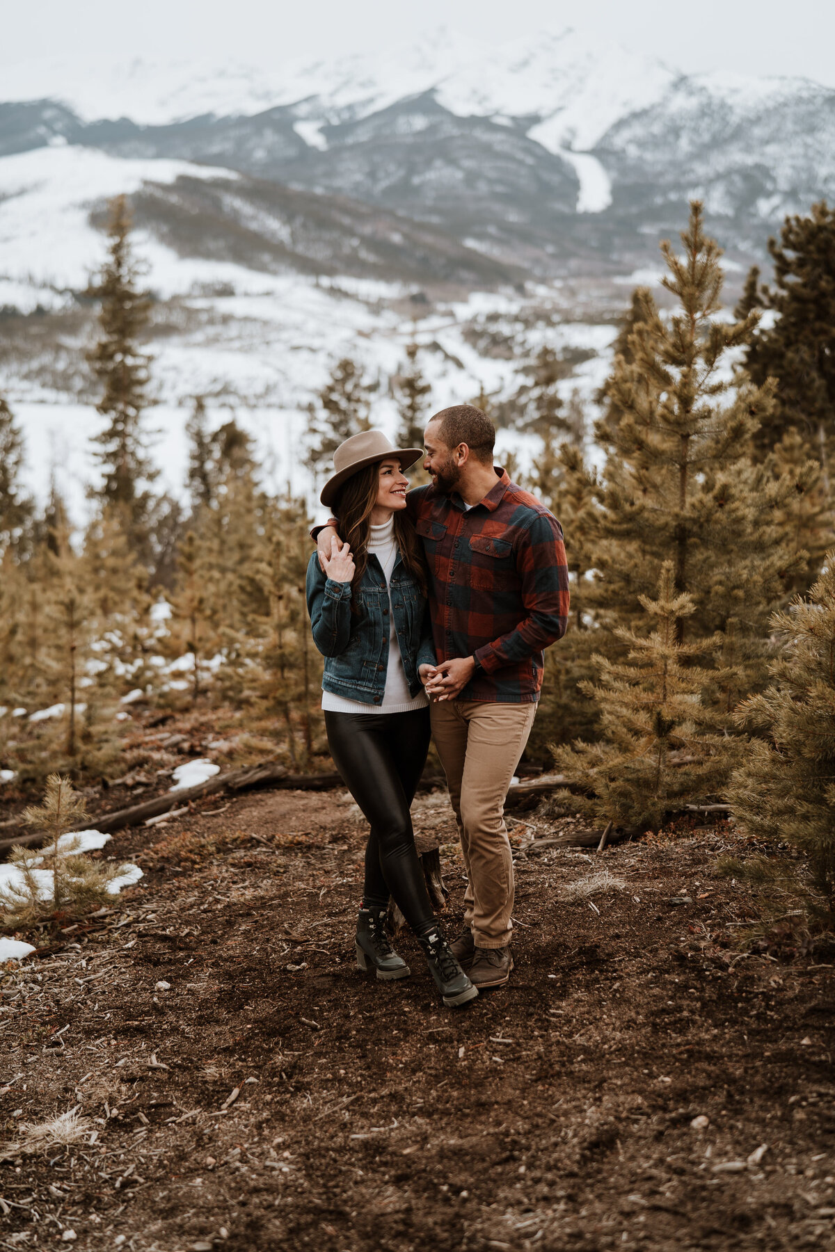 Engaged couple walks through pine forest near Breckenridge with mountains in background.
