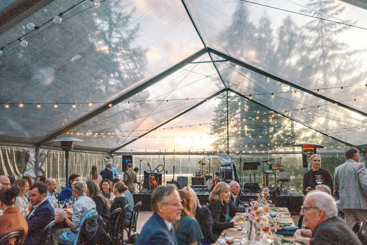 The inside of a clear reception tent with people seated at tables and trees visible in the background.
