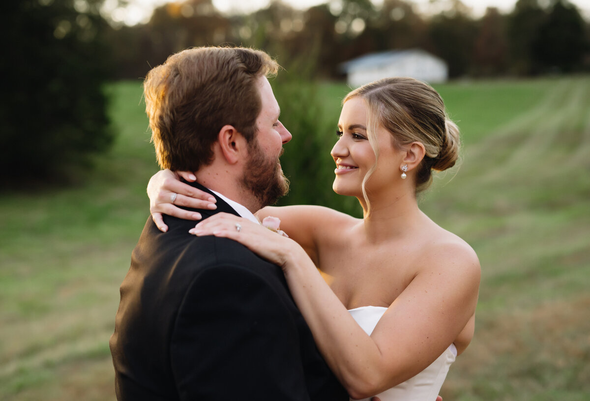 bride and groom embracing on a hill with a tree line in the distance on a green hill while the sunshines onto the couple as it sets in the distance
