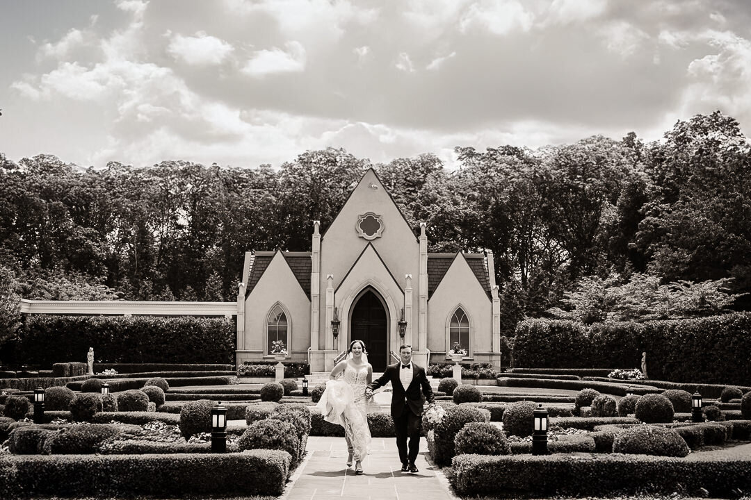 Bride and groom walk hand in hand away from a beautiful outdoor chapel after their wedding ceremony, captured in black and white.