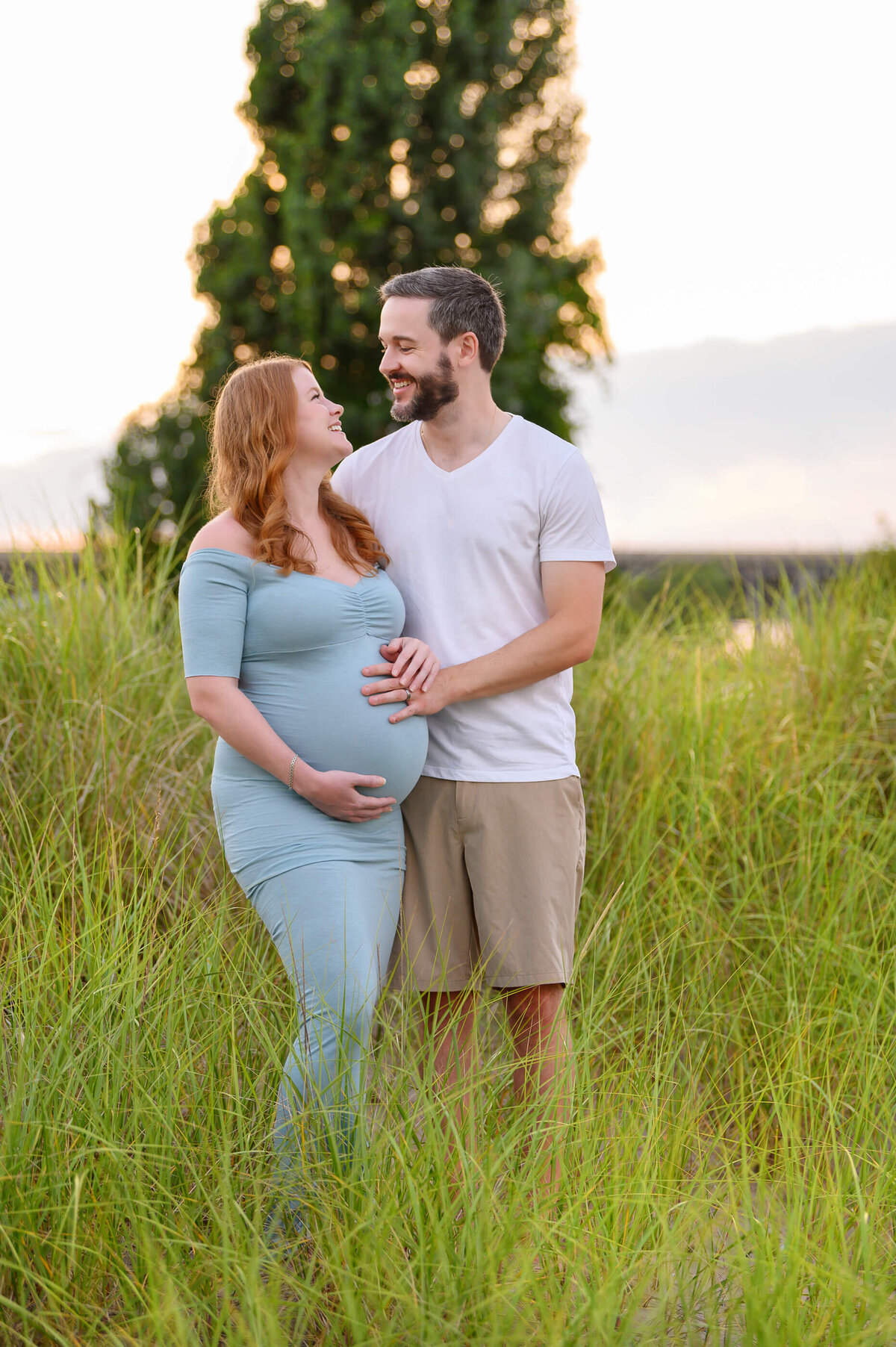 husband and wife standing cradling mom's belly in tall grass for her sunset beach maternity photos toronto