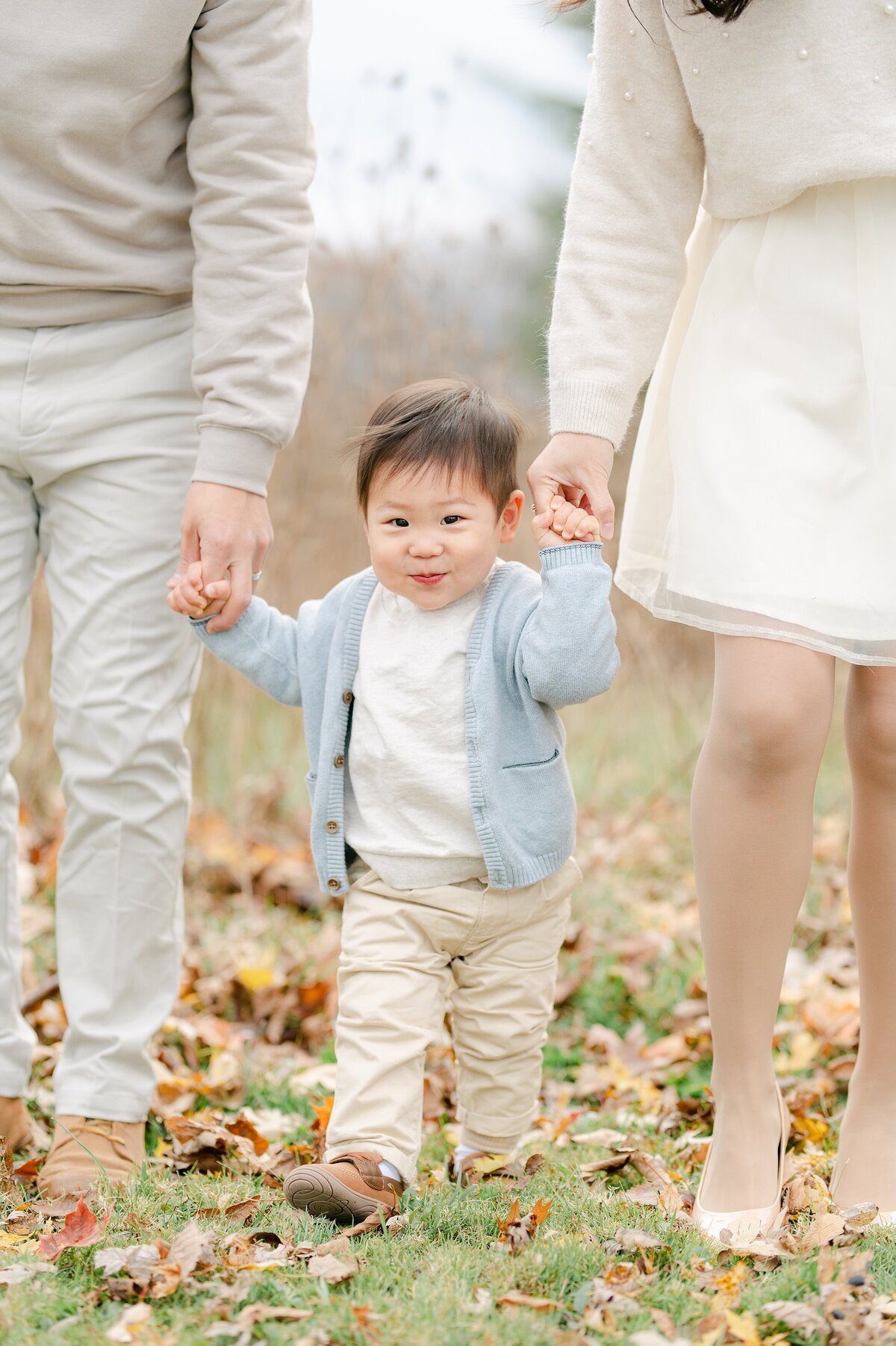 Little boy walks through leaves while holding parents' hands and smiling