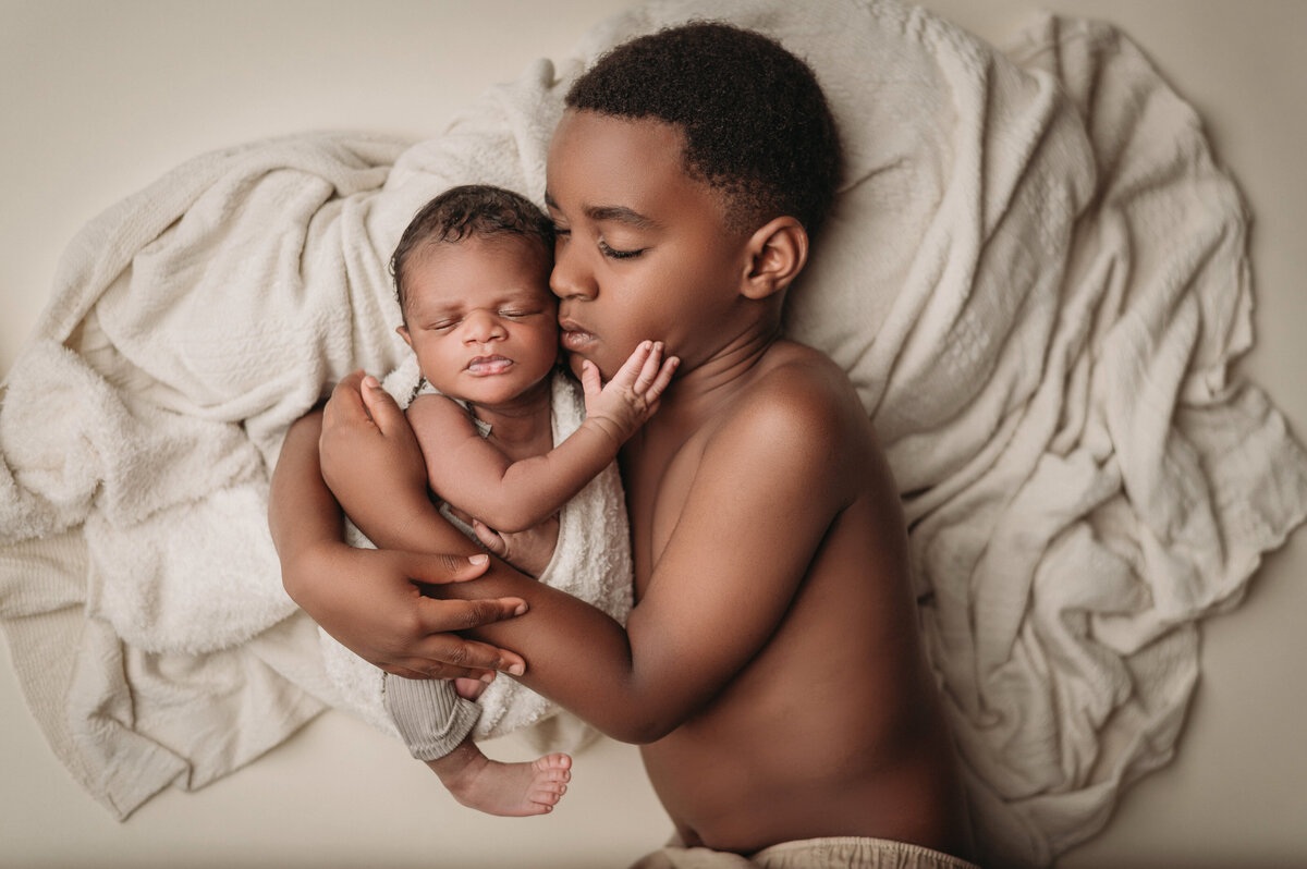 sleeping big brother  snuggling close his new baby brother wrapped in cloth while baby holds  his brother's face with his hand and they are over a blanket and cream backdrop