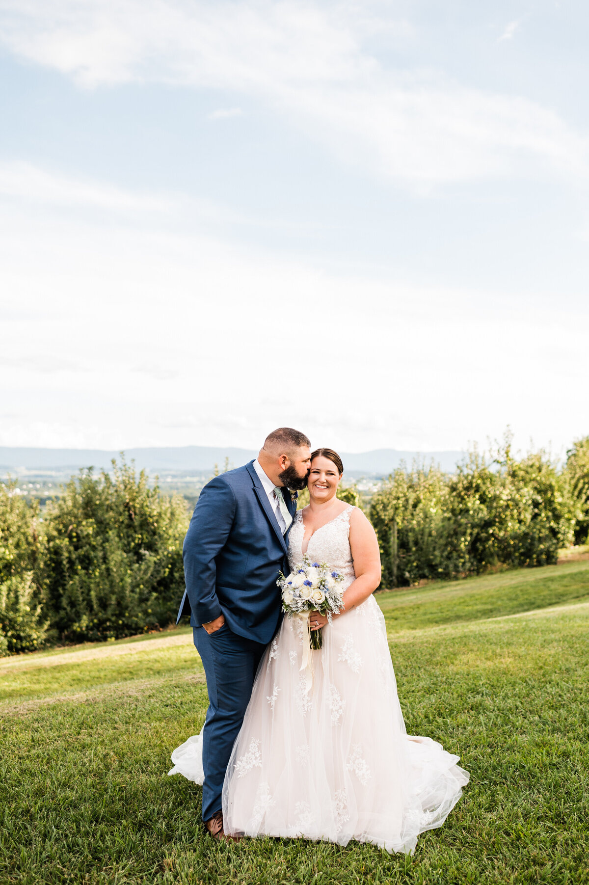 Virginia wedding photographer captures bridal portrait of bride holding her white floralsas her groom kisses her cheek with the mountain range in the distance