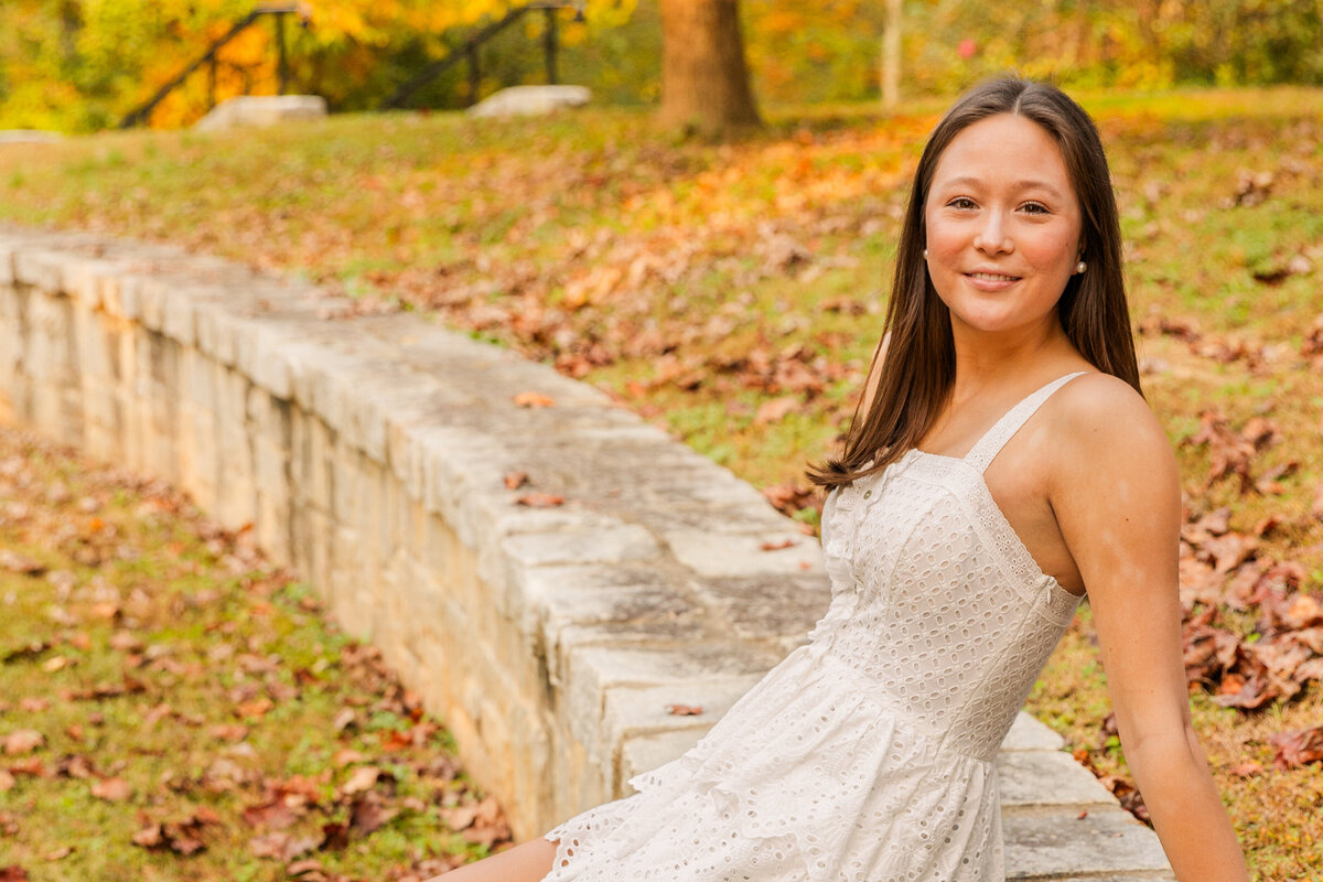 wide shot of senior girl siting in a Atlanta park wearing a short white dress during senior photo session