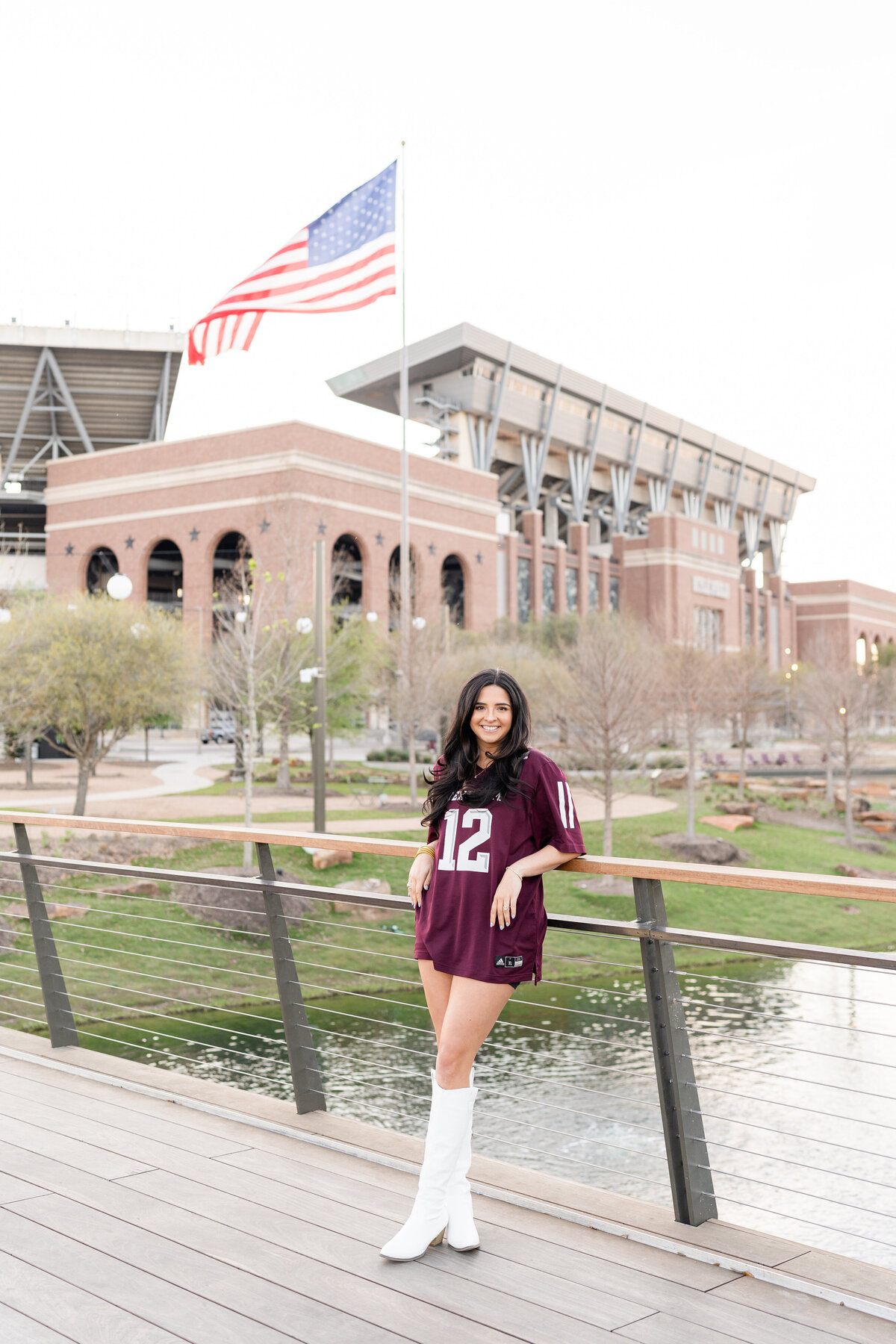 Texas A&M senior girl leaning against bridge railing with Kyle Field in the background while wearing white boots and maroon Aggie jersey