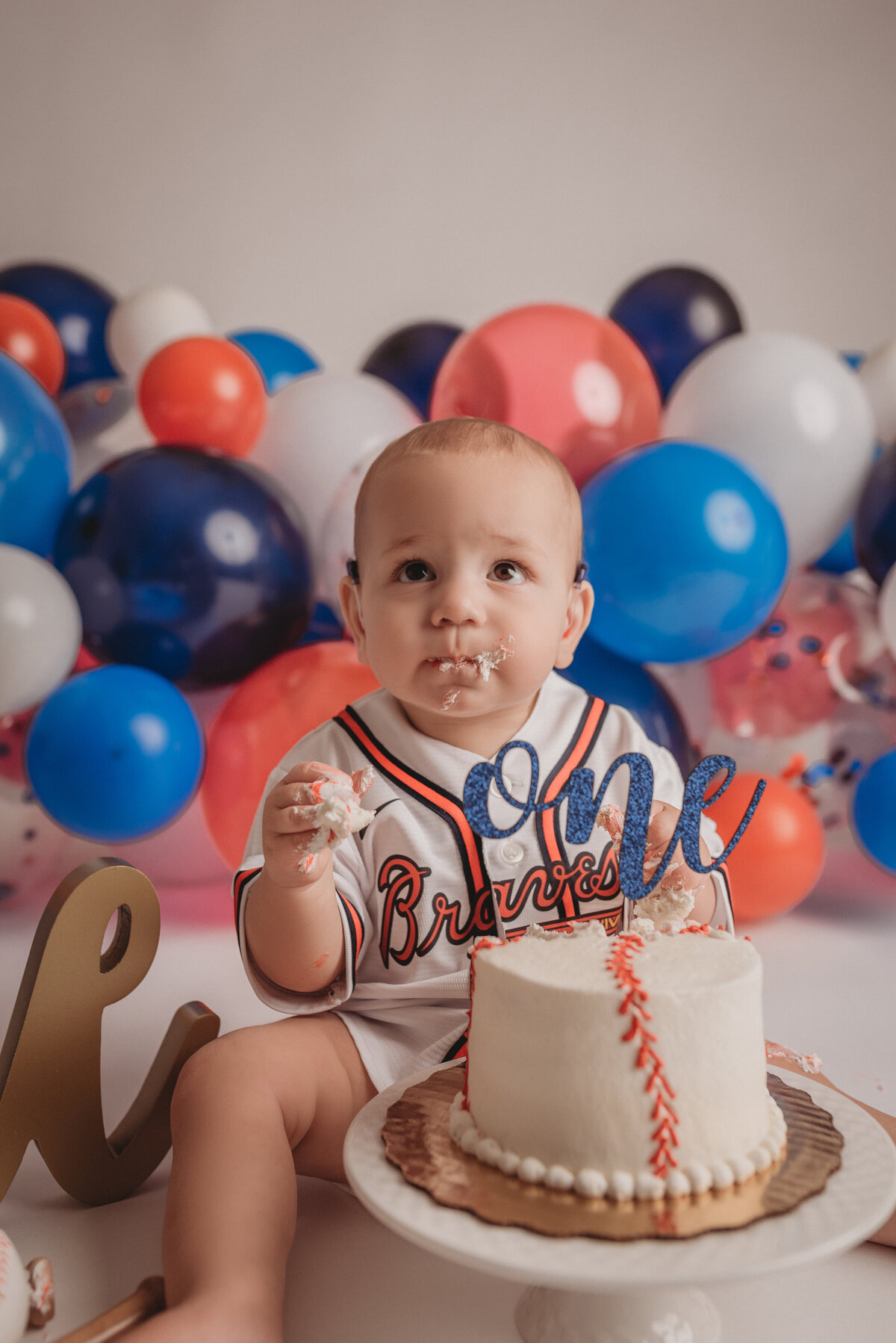 One year old baby boy sitting on floor eating birthday cake with Atlanta Braves baseball jersey on and red, white, blue balloon garland behind him