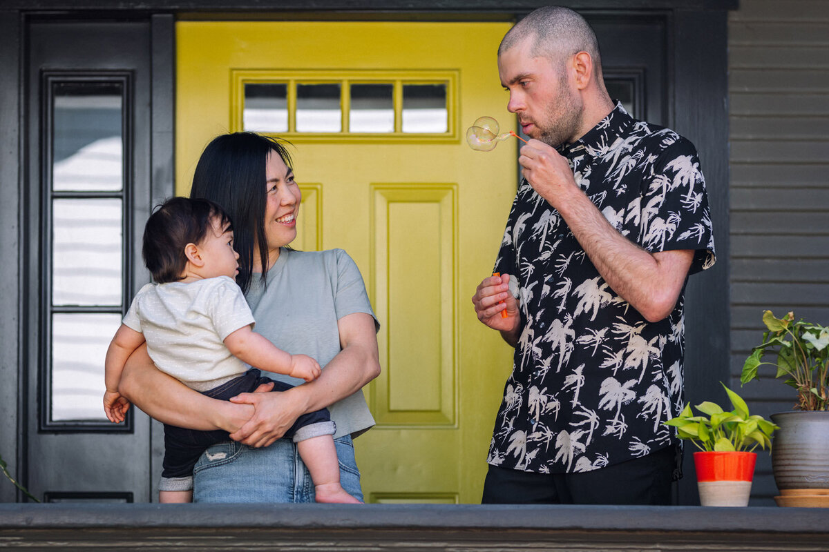 parents and toddler girl standing on front porch with yellow door and dad is blowing bubbles to baby in mom's arms