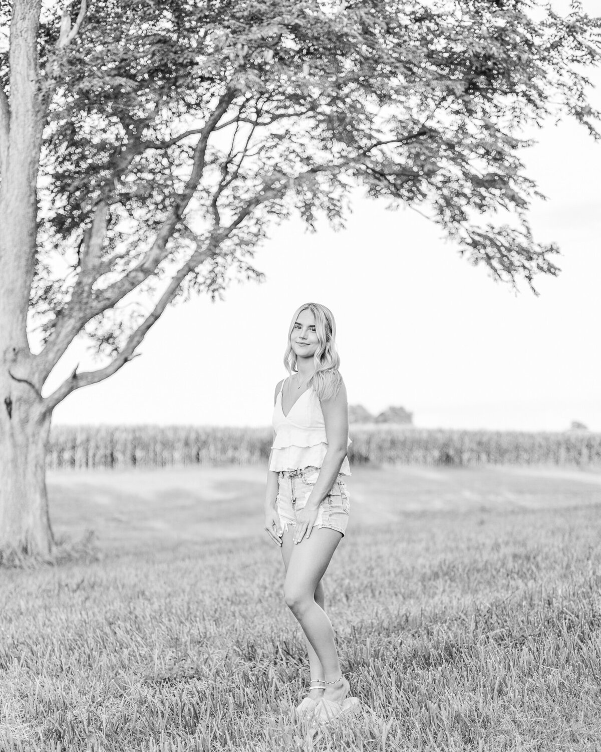 black and white photo of a high school senior female with long hair, standing in a field with a tree and cornfield in the back, turned to the side, with a soft smile at the camera, wearing blue jean shorts, and a spaghetti strapped shirt