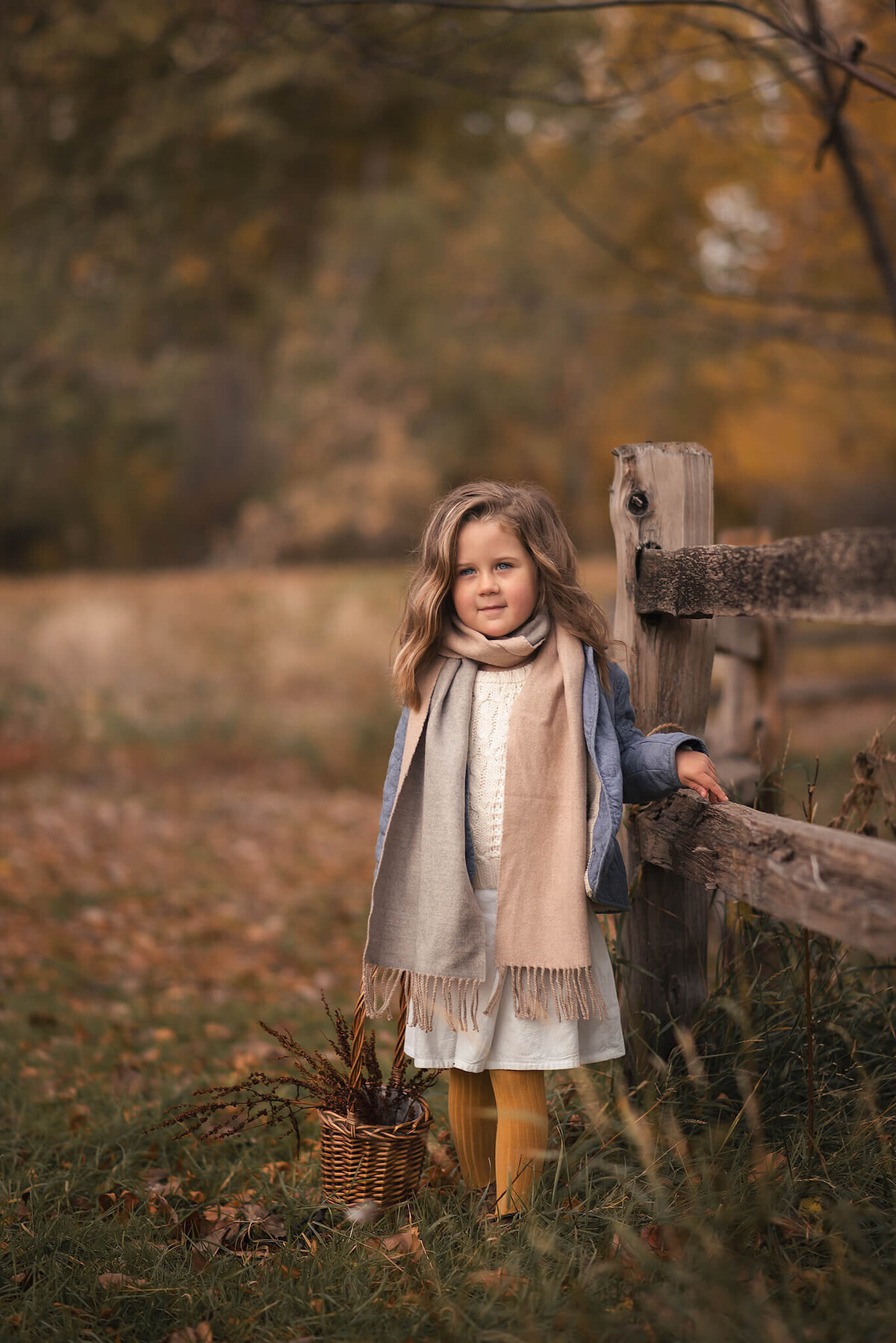 A little girl is dressed in fall layers, leaning against a fence while smilling at the camera with a grogeous background in Kamloops.