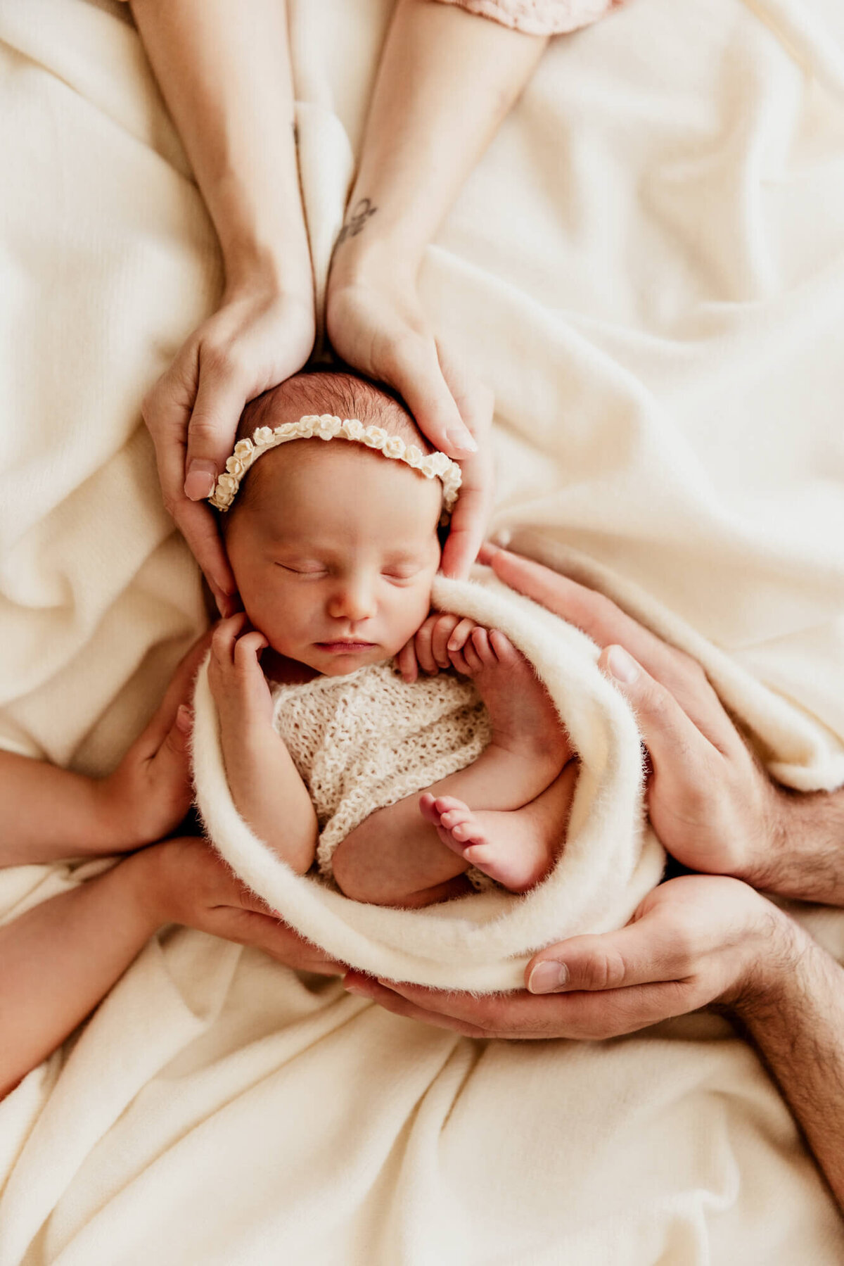 Family of three places hands on their daughter who is sleeping in a swaddle on a white blanket.
