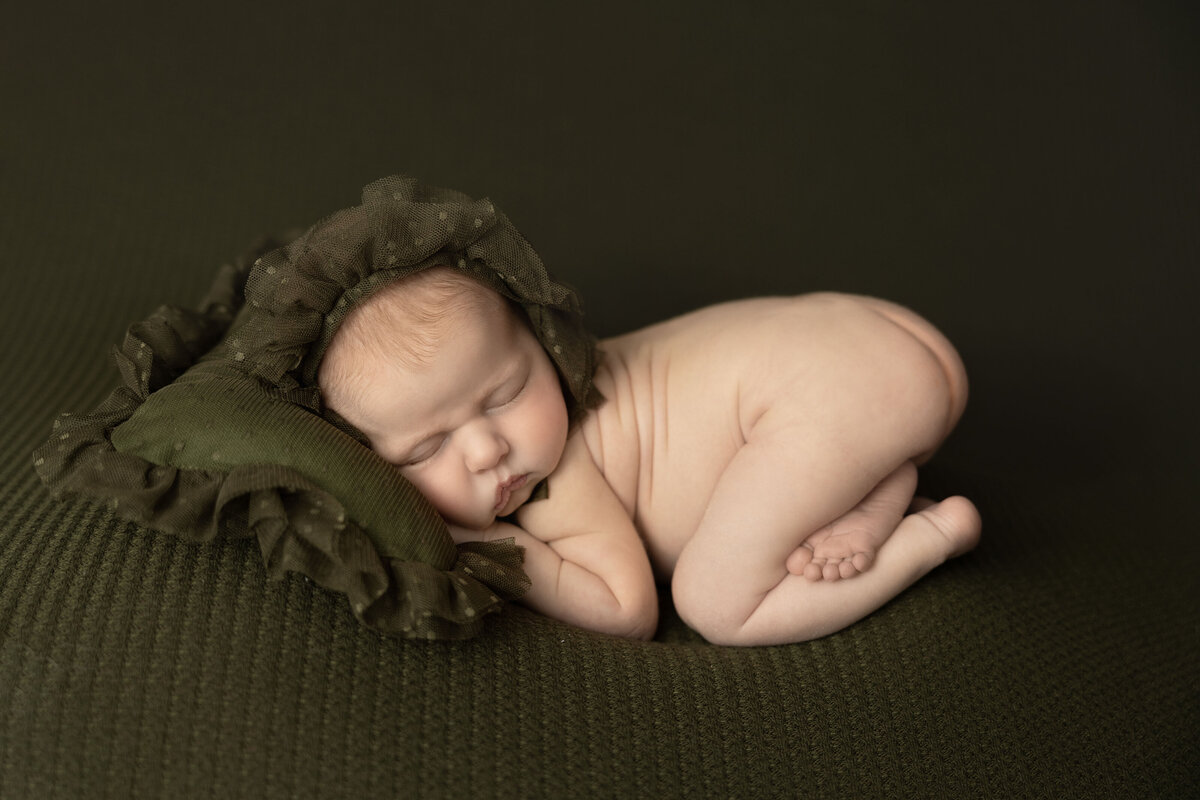 A newborn  dressed in a green bonnet, snuggles a frilly green pillow in a St. Louis photography studio.