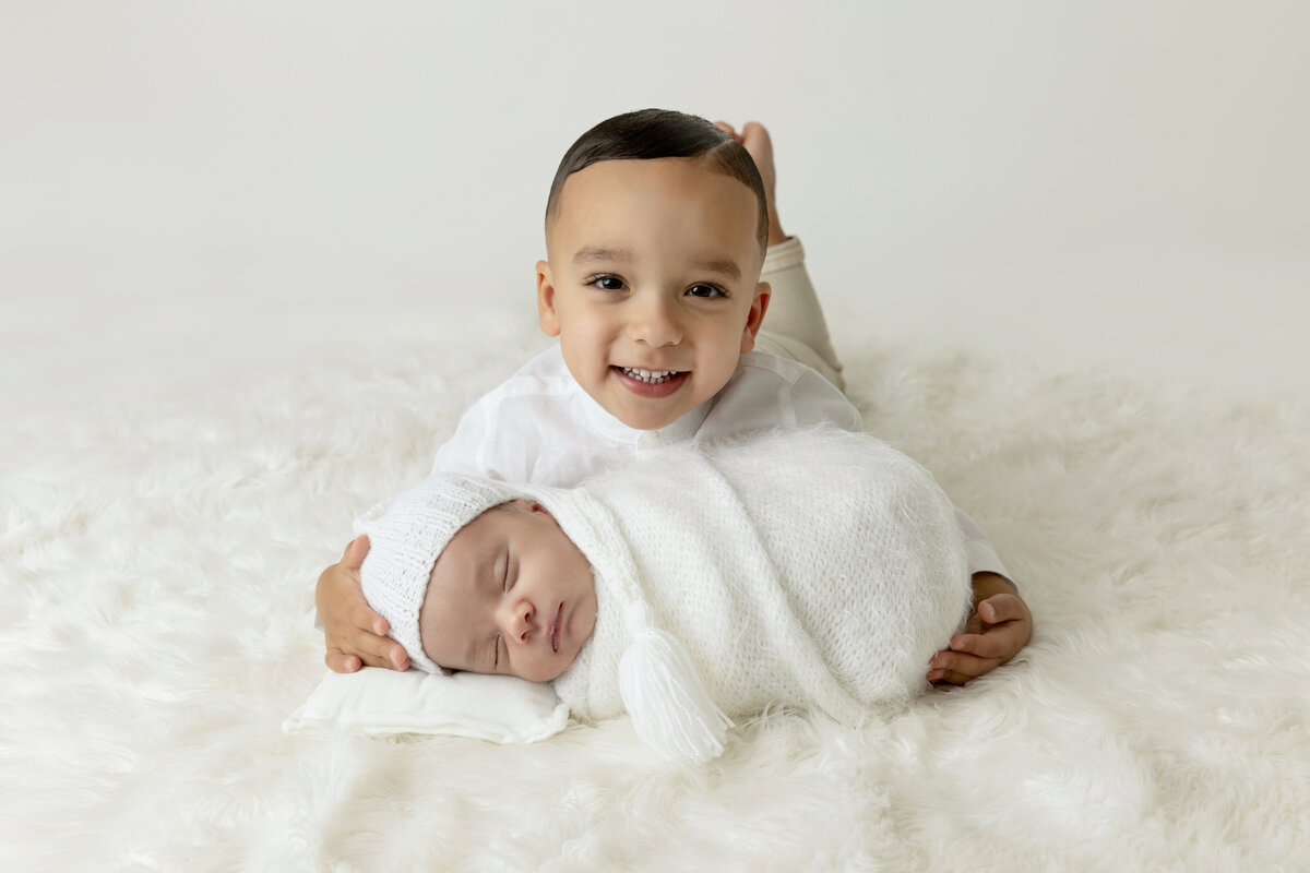 A happy toddler bow lays on a bed holding his sleeping newborn baby sibling in front of him in a white swaddle and hat