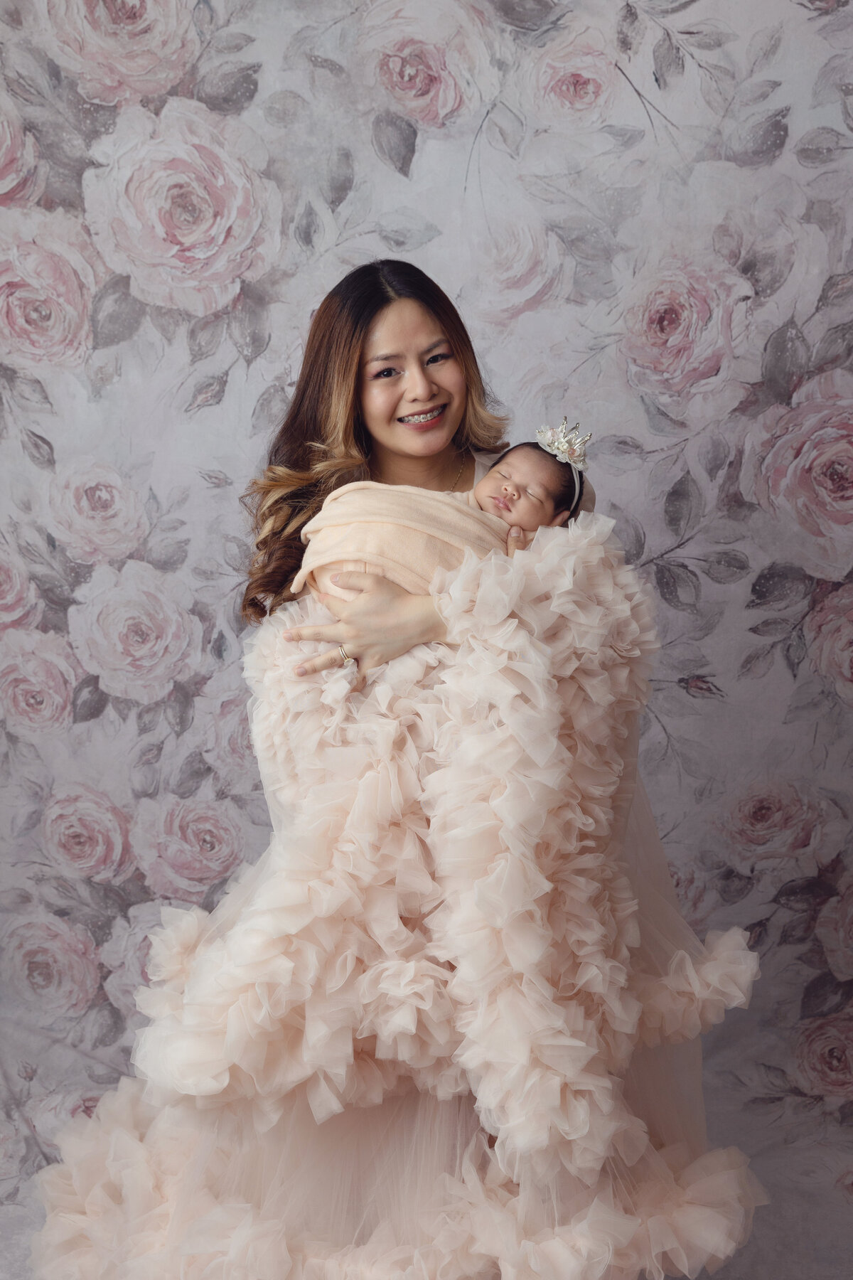 A mother in a flowing tule dress smiles with her sleeping newborn baby daughter in her arms in a matching swaddle