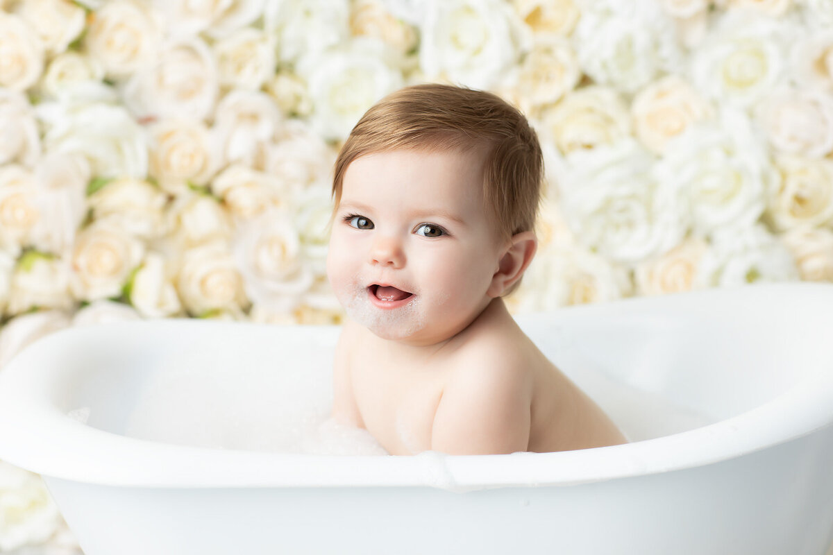 Milestone baby session by a Dallas milestone photographer featuring a smiling baby in a bubble bath against a backdrop of ivory roses, capturing a playful and tender post-cake smash moment.