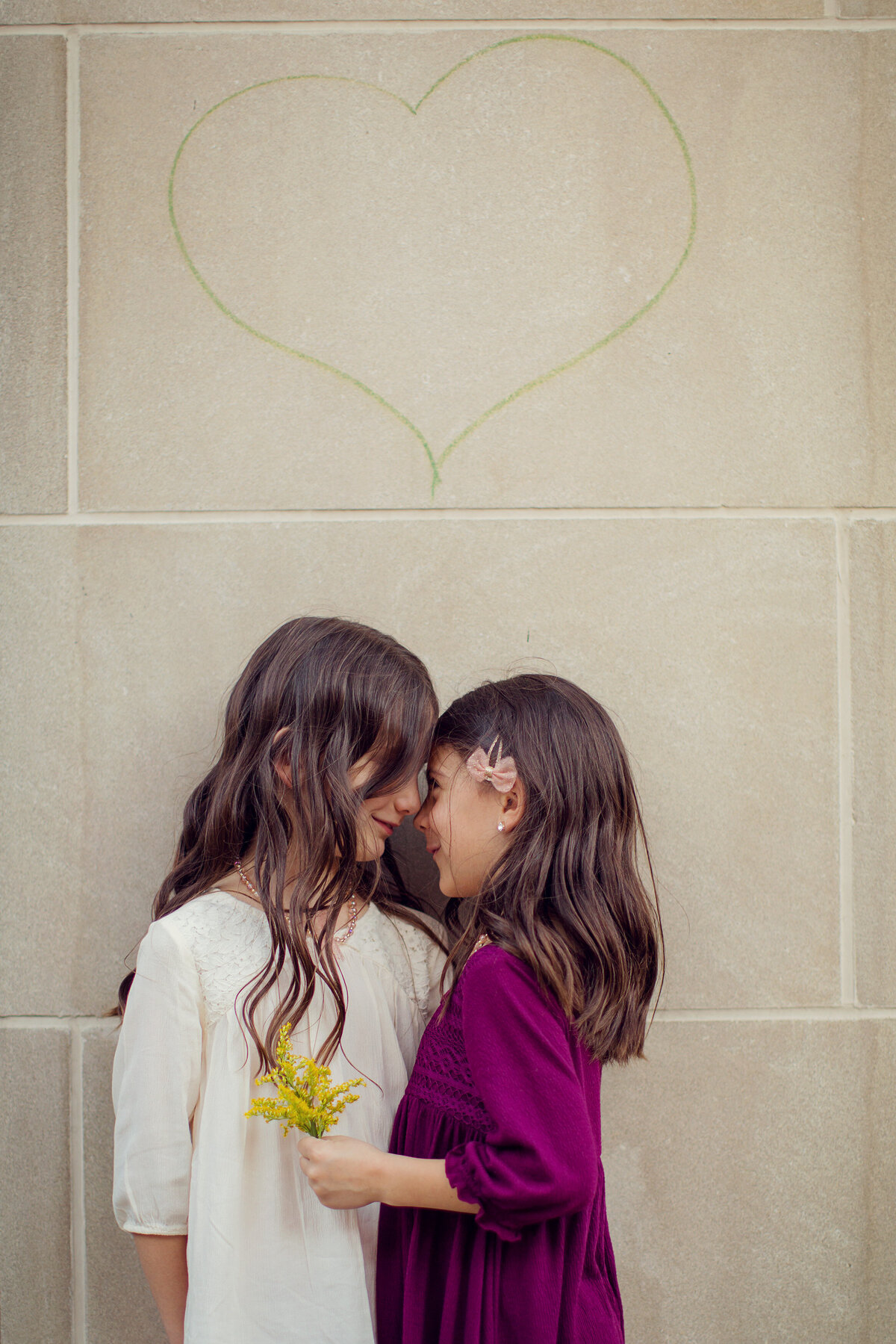 Two sisters in dresses are face to face in downtown Indy against a concrete wall that has a green heart on it.
