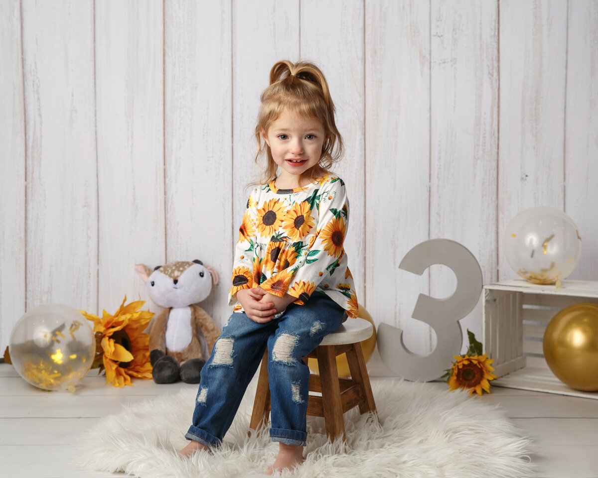 Cute three year old girl wearing a sunflower shirt and jeans, sitting on a stool with balloons and the number three in the background