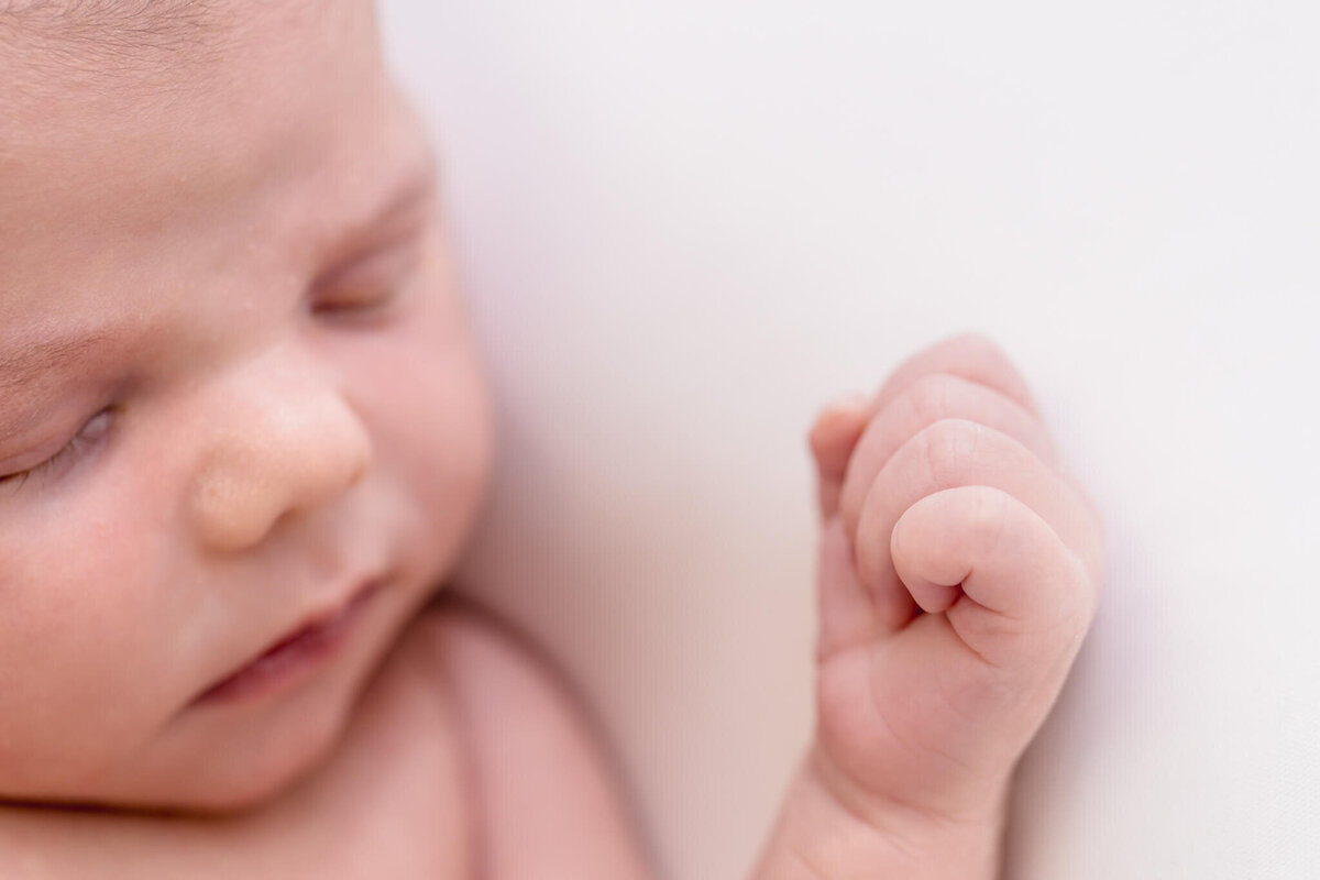 Close-up of a sleeping baby with a focus on the babys closed eyes and small hand clenched into a fist, resting gently against a soft white background.
