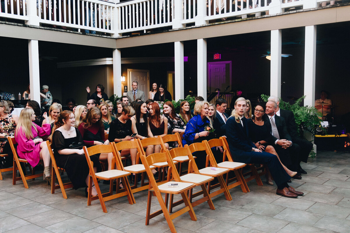 A group of people dressed formally are seated in wooden chairs at an outdoor wedding venue, some chatting and smiling, with empty chairs in the front and a balcony above.