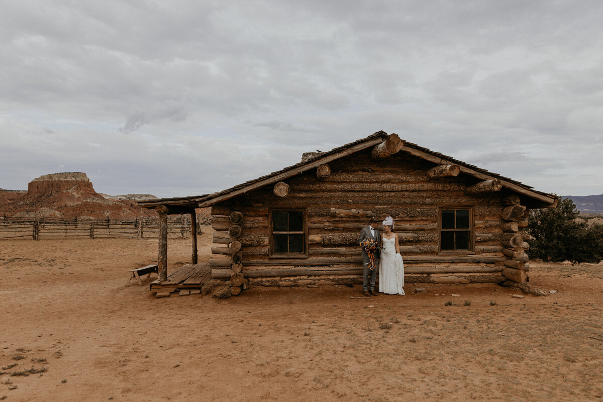 bride and groom standing outside of an old log cabin in the desert