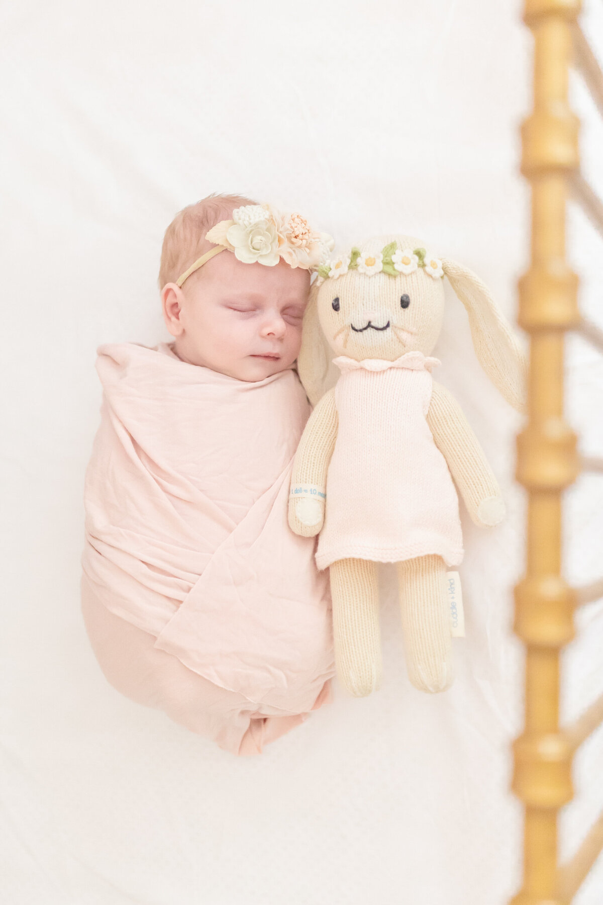 Newborn baby girl swaddled in her crib, resting peacefully beside an adorable little stuffed doll. The soft, gorgeous light highlights her sweet and delicate features, creating a warm and serene atmosphere
