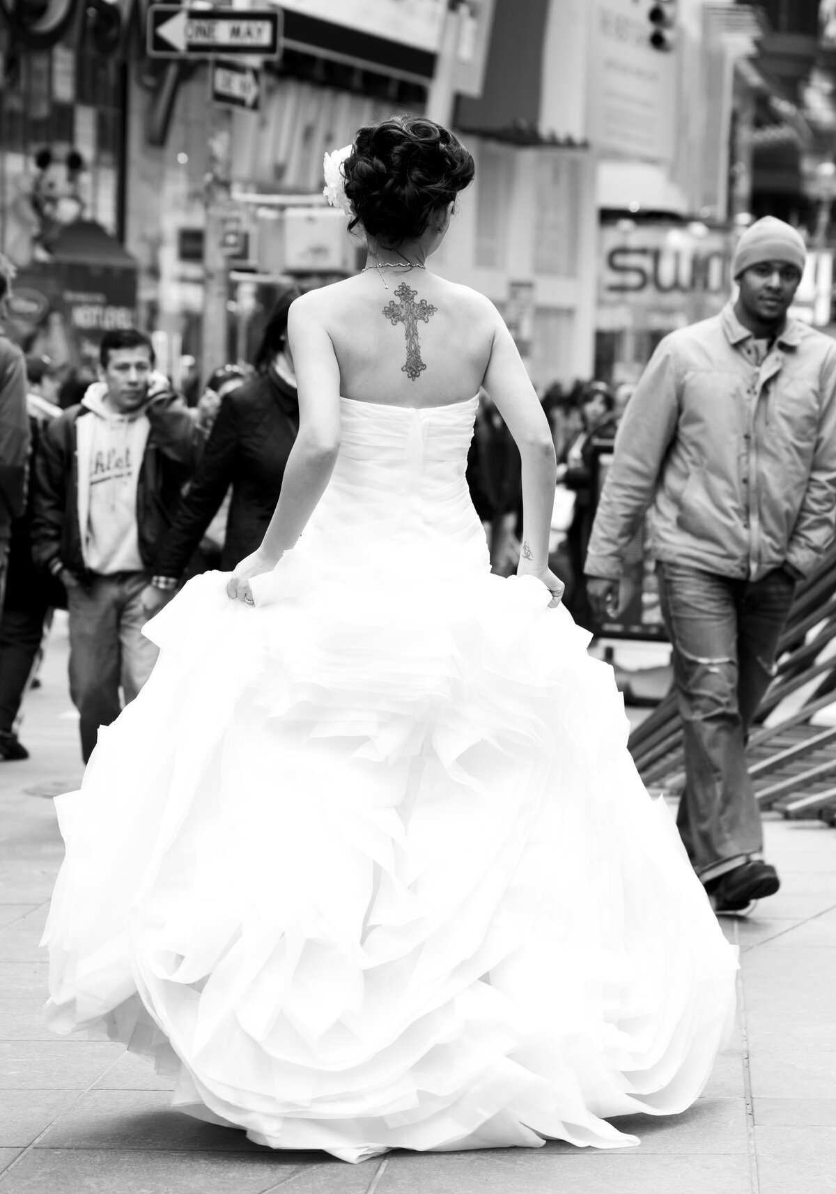 A black and white photograph capturing a bride in a voluminous wedding gown standing  in Times Square, New York City. her back is to  the camera, showcasing a detailed cross tattoo on her upper bak. Her hair is styled elegantly in an updo with a flower accessory.  the bustling city life continues around her, with pedestrians in various attire walking by, unfazed by the bride's presence.