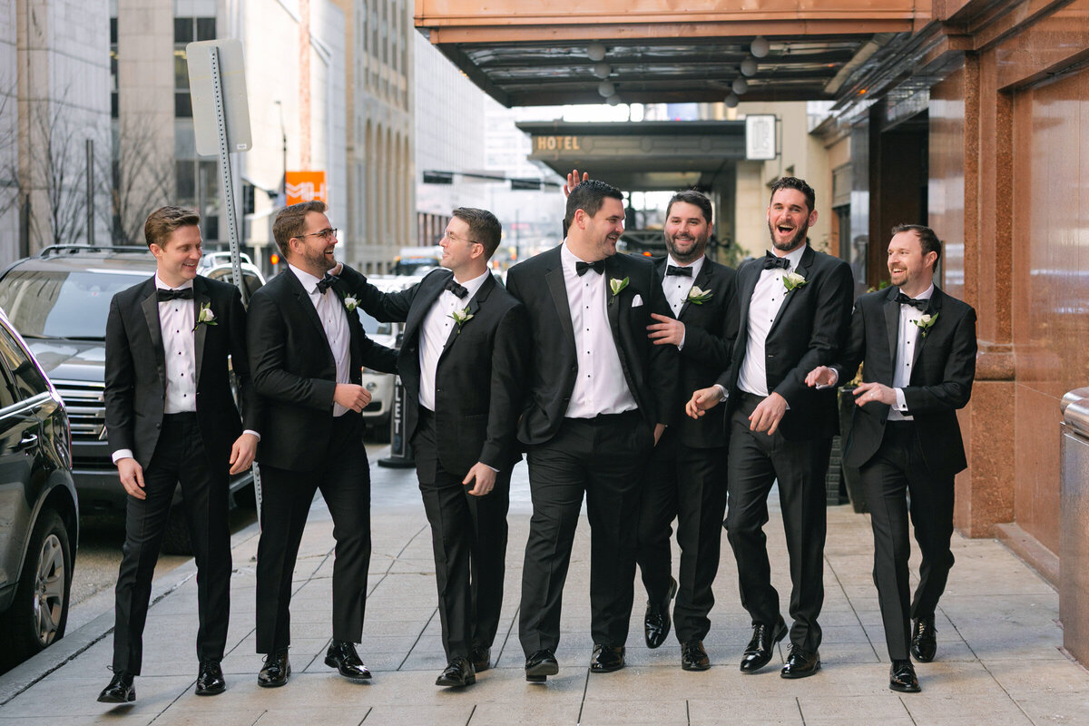 groom and groomsmen laughing while walking outside the entrance of The Adolphus in Dallas, capturing a joyful and candid wedding moment.