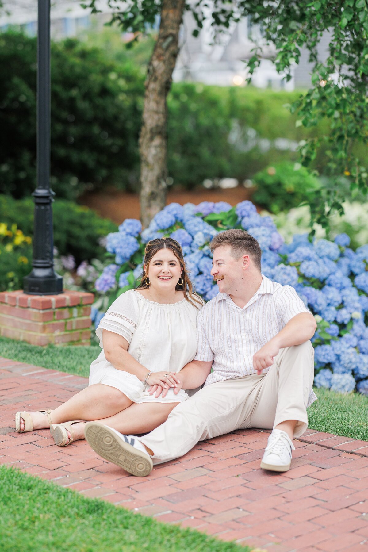 Couple sitting on a brick walkway, holding hands and smiling at each other, with vibrant blue hydrangeas and green foliage in the background at Chatham Bars Inn in Chatham, Massachusetts — representing the portfolio of a Cape Cod engagement photographer.
