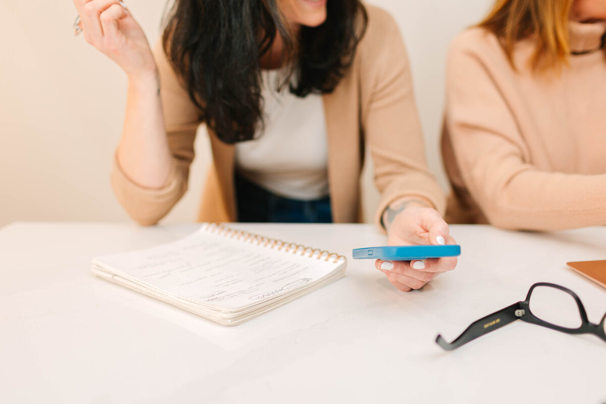 A woman in a beige cardigan holds a blue smartphone while discussing notes in a spiral-bound planner. Another woman, also in neutral tones, sits beside her, with black glasses resting on the white table.