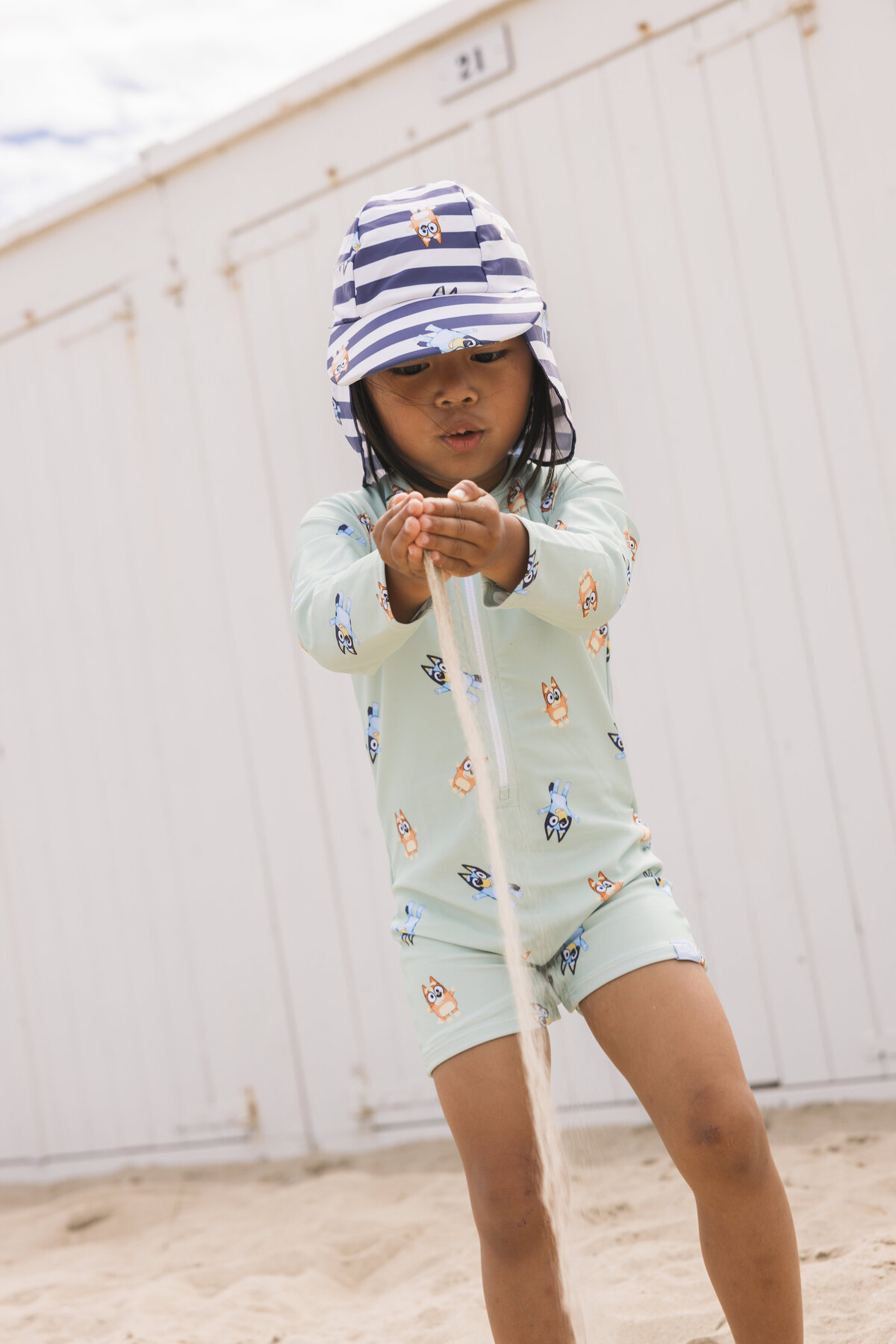 Child playing with sand at the beach, wearing a mint green Swim Essentials UV suit and a striped sun hat.