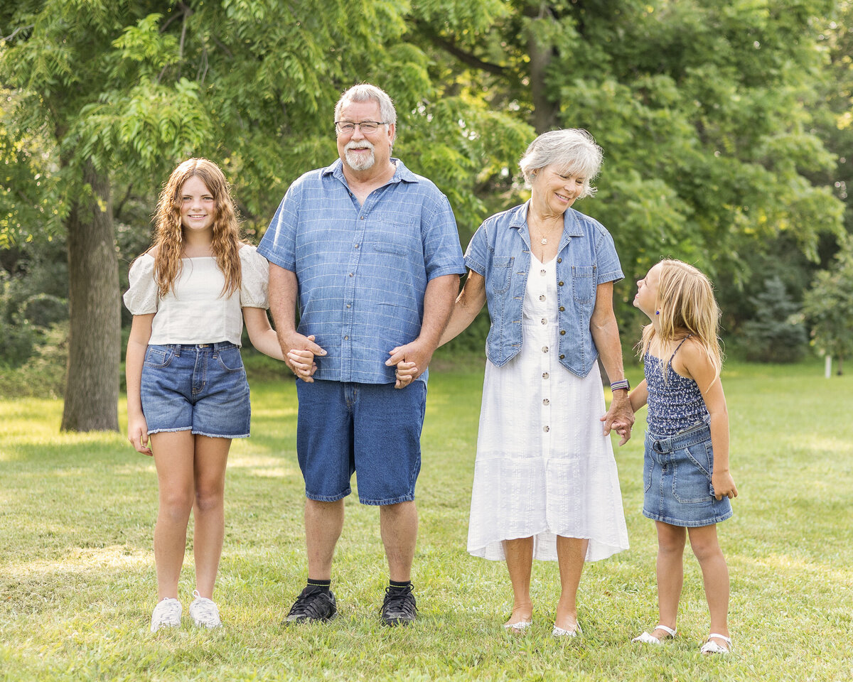 Grandparents and granddaughters standing next to each other, holding hands, in grass in front of a tree line, smiling at each other and the camera.  Older daughter is wearing blue jean shorts, a whitle short sleeve shirt, and has long brown hair.  Grandpa is wearing a light blue and white stripped short sleeve button up shirt, blue jean shorts, and has white short hair and goatee.  Grandma is wearing a white button up dress with a jean jacket over it, with short white hair.  Youngest daughter has a dark blue and white tank top style shirt with a blue jean skirt, and has medium length blonde hair.