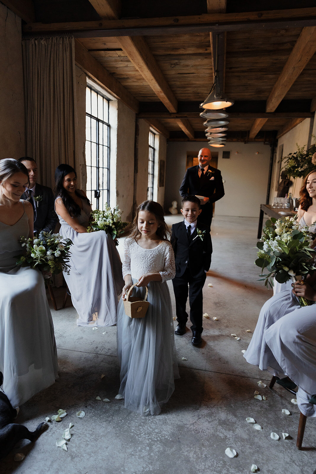 flower girl walks down aisle dropping roses with the ring bearer behind her and the groom coming last