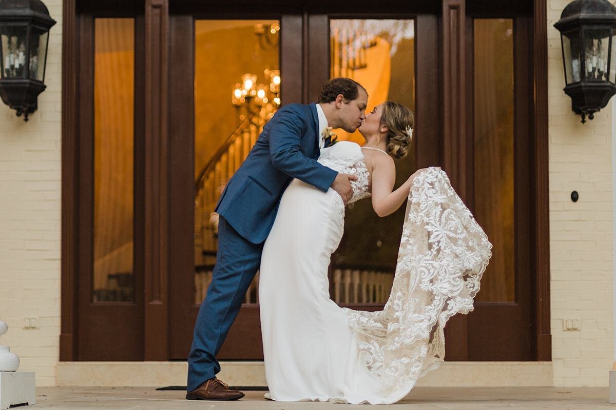 The groom, wearing a blue suit and brown shoes, dips the bride, wearing a strapless lace wedding dress with a long lace train, backwards as he kisses her in front of a set of glass doors in dark wood door frames with a brass chandelier  and spiral staircase behind them. On either side of the Estate at Cherokee Dock there are two large black wall sconces on the light beige brick walls. Nashville Wedding.