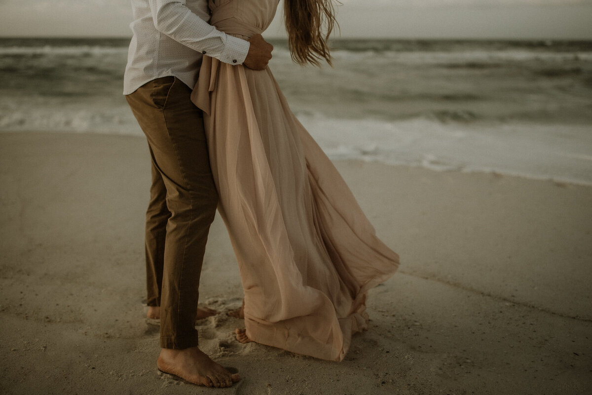 Couple on a beach, barefoot in the sand with the wind blowing through her hair and dress.