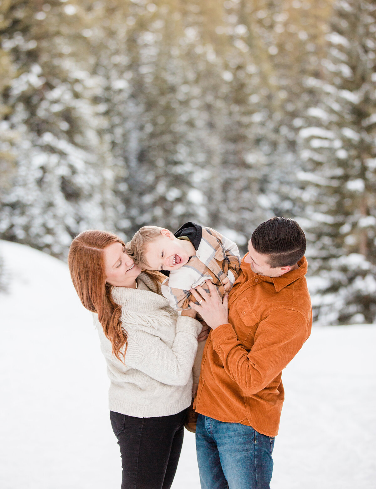 Parents have their young son in their arms and are tickling him playfully. There is a scene of snow covered pine trees behind then