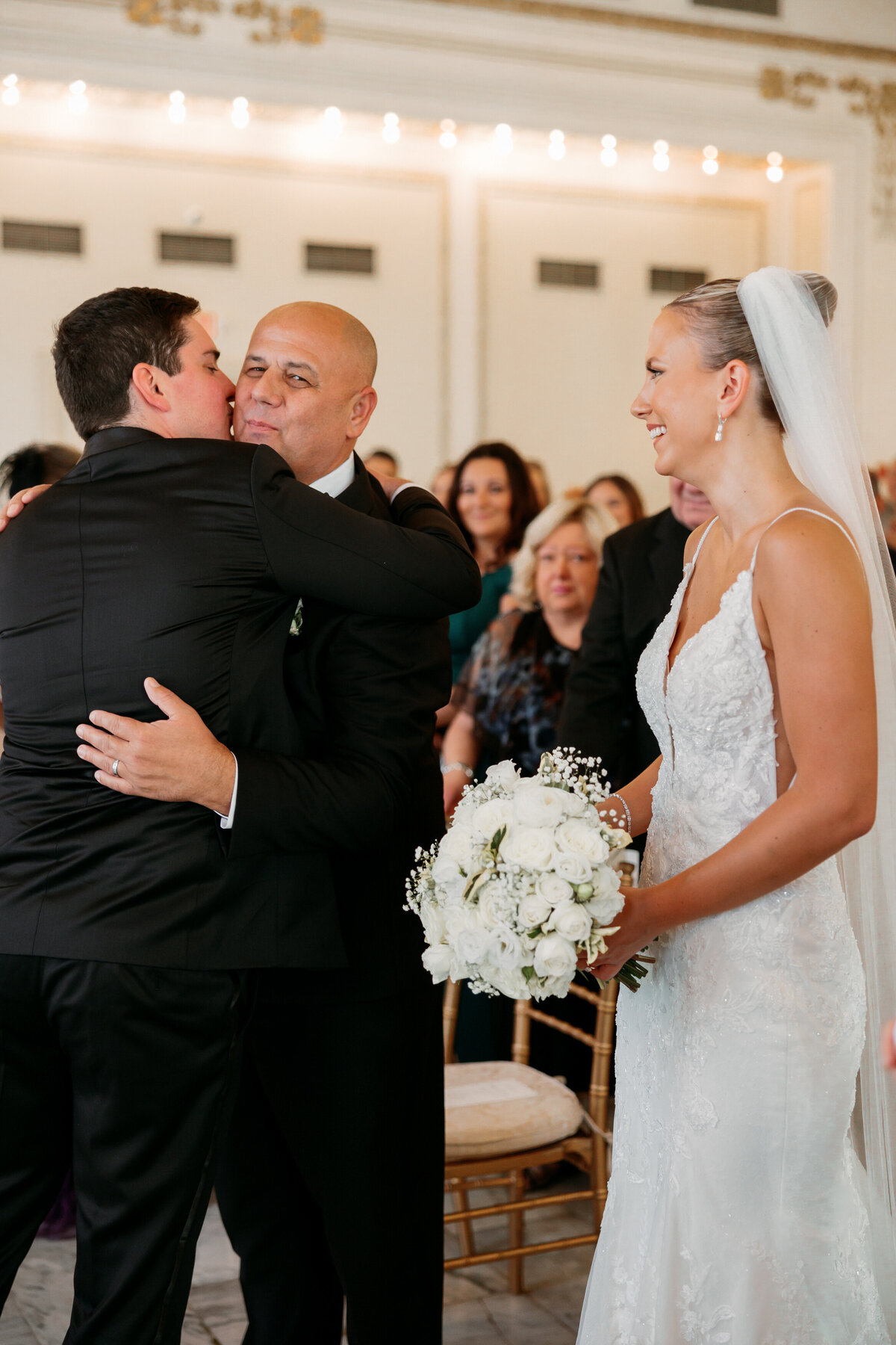 Groom hugs the bride’s father while she looks on smiling, holding her white rose bouquet—intimate ceremony detail from a Philadelphia wedding photographer.