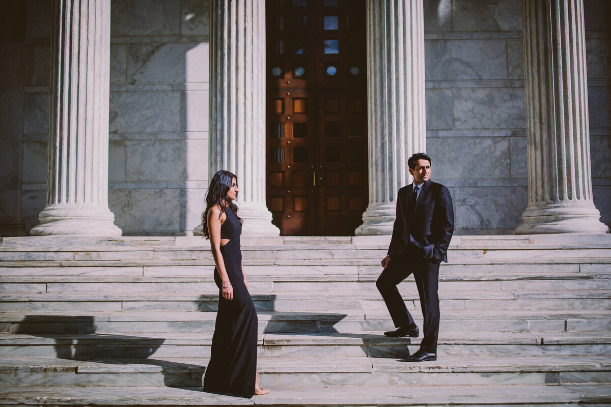 A couple standing in black formal outfits separetely on the steps of the clio whig hall building at princeton university in nj during their engagement session