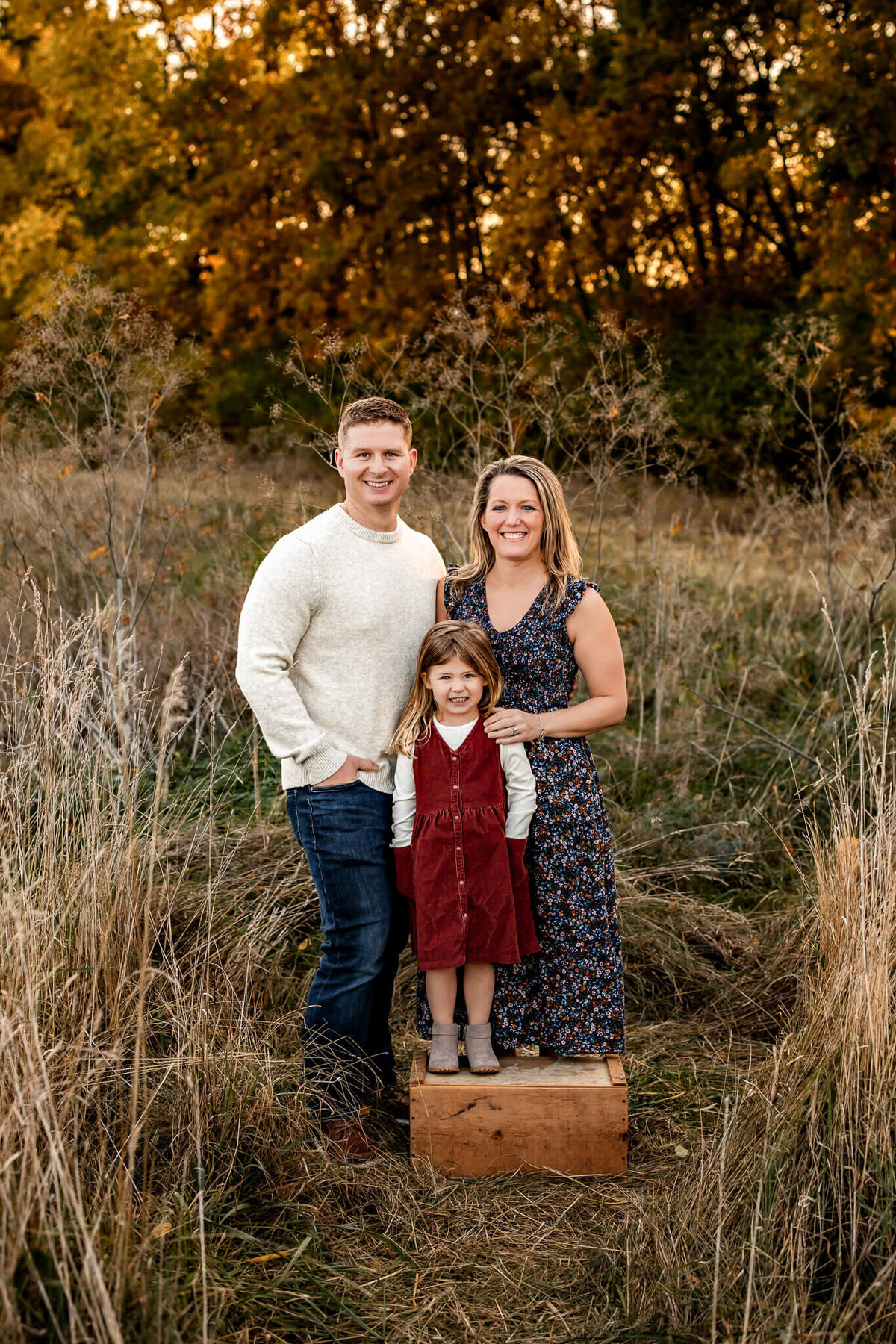 A Bloomington family photographer captures a charming outdoor portrait: a family of three amid autumn foliage. The child stands on a wooden box in a red dress, flanked by smiling parents in casual attire, all surrounded by tall grass.