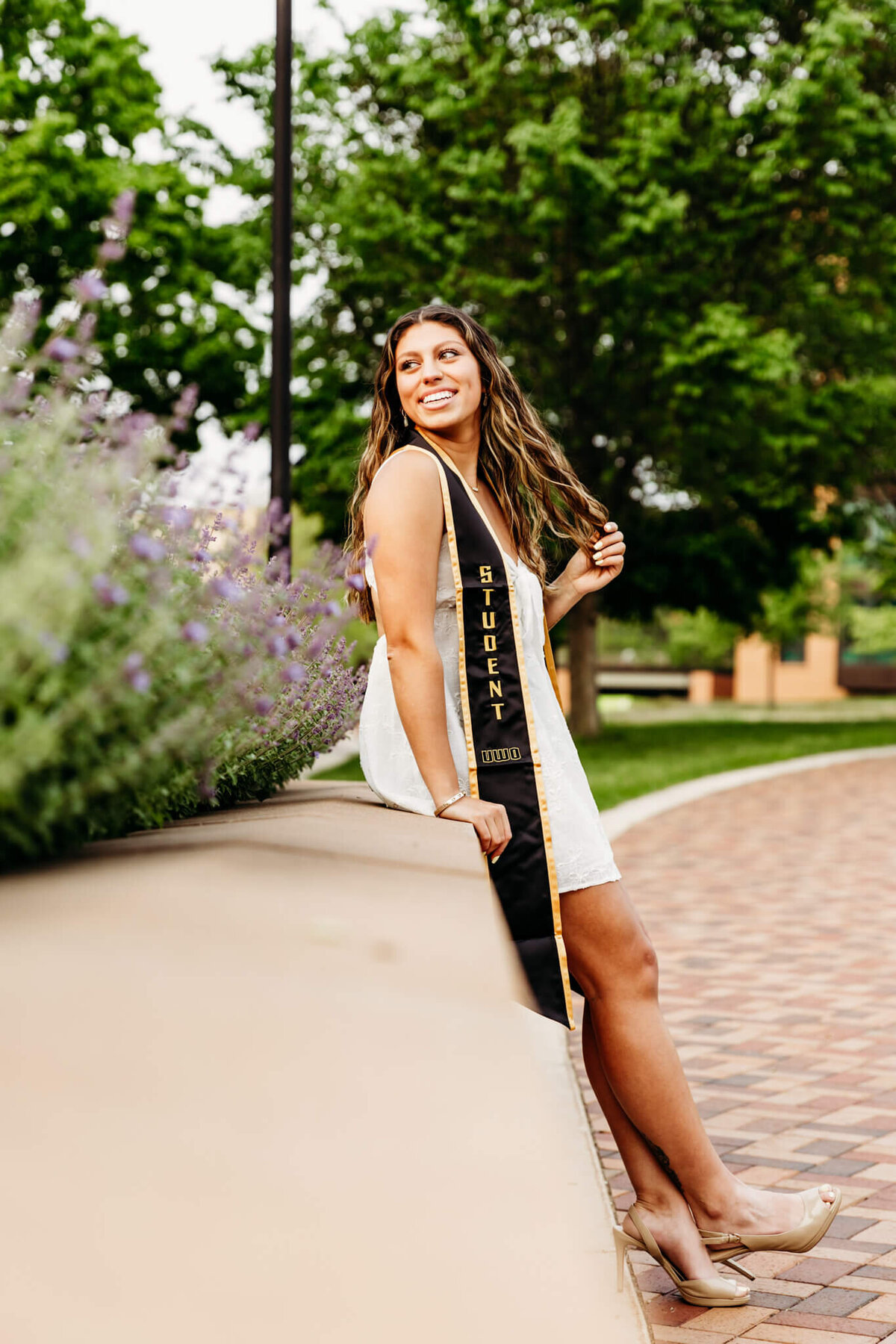 Gorgeous UW Oshkosh graduate playing with her hair and she leans against a ledge and looks over her shoulder at the purple flowers captured by Oshkosh Graduation Photographer Ashley Kalbus