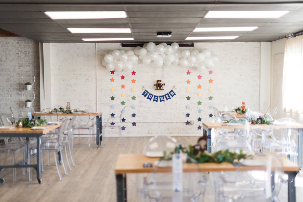 Whimsical backdrop for a baby boy shower, set against a white brick wall, with rainbow star garland, white balloon clouds, and a teddy bear sitting on a wooden swing, with the baby's name displayed in fabric garland below.