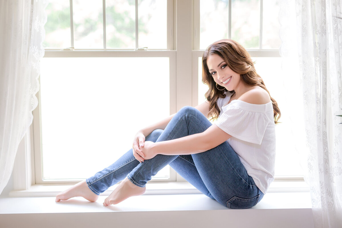 Female headshot client sitting on a windowsill smiling at the camera wearing jeans & a t-shirt