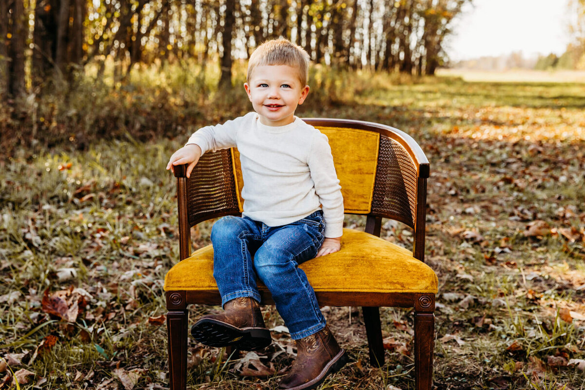 Young boy sitting in a yellow velvet chair in a field surrounded by trees by Milwaukee Family Photographer Ashley Kalbus.