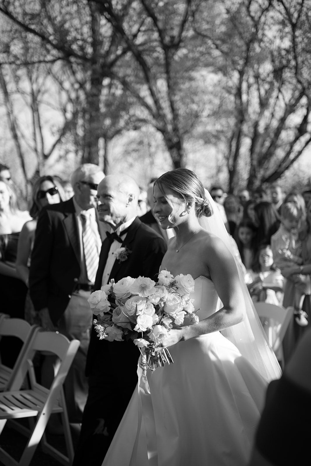 Black and white photo of the bride holding her bouquet and smiling as she walks arm-in-arm with her father down the aisle during the wedding ceremony at Burritt on the Mountain in Huntsville, Alabama, photographed by Perry Hancock.