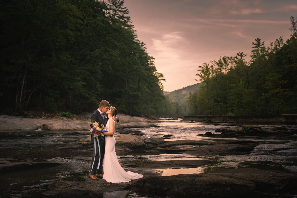 Bride and groom standing on river rocks with forest and mountains in the background, serene sunset landscape