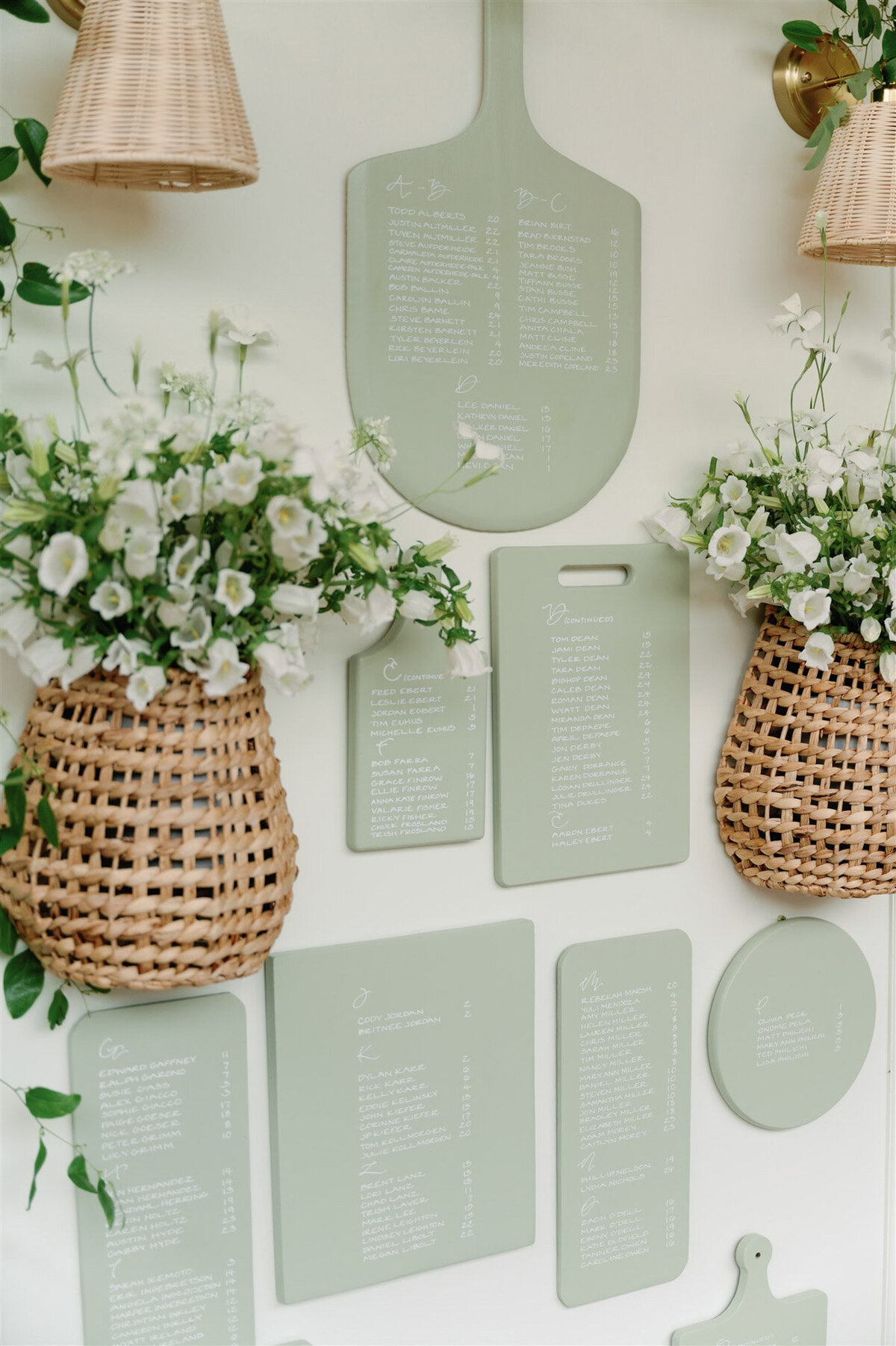Green cutting boards with custom calligraphy details for a seating chart hanging on a wall with florals, accent lights and baskets representing full service design and calligraphy by Life in Letters.