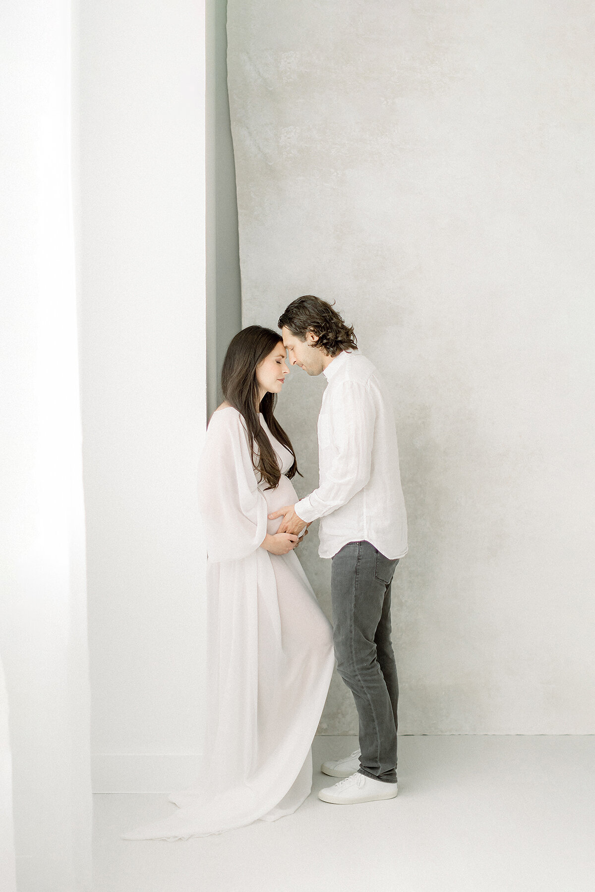 Photo of an expecting Dallas mom leaning on a wall with her spouse standing in front of her as he holder her belly for their maternity session at a Dallas photography studio.