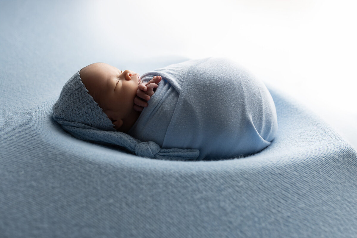 Side view of swaddled baby in blue with light streaming in, captured by Dallas newborn photography studio.