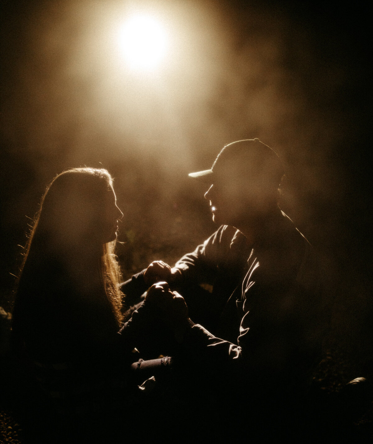 Silhouetted couple sitting on the ground straddling each other in front of a motorcycle headlight on a foggy night